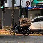 Motociclista profissional com caixas empilhadas aguarda o sinal abrir em uma rua movimentada da cidade.