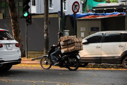Motociclista profissional com caixas empilhadas aguarda o sinal abrir em uma rua movimentada da cidade.