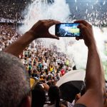 Torcedores nas arquibancadas filmam uma partida de futebol com um smartphone.