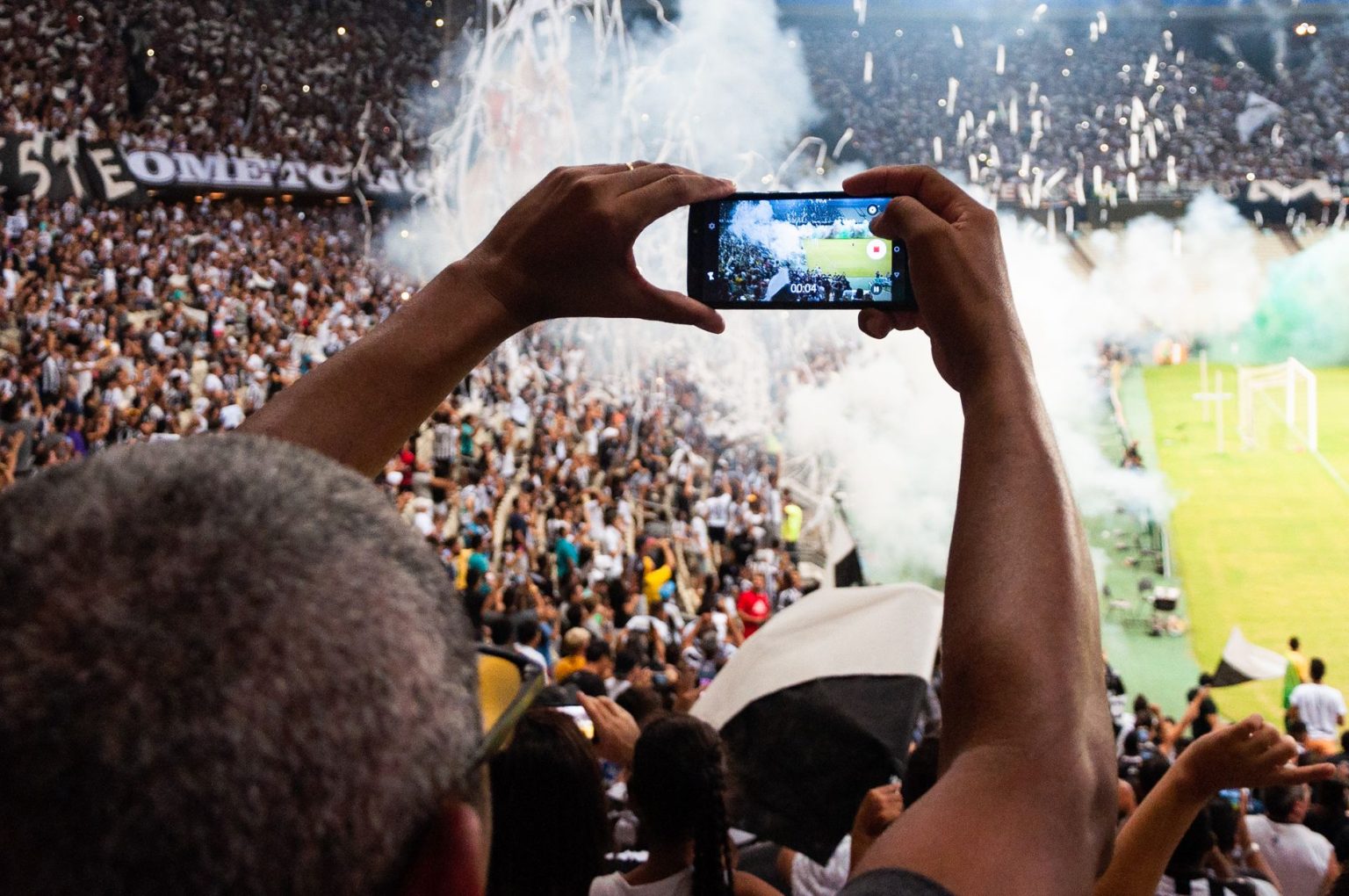 Torcedores nas arquibancadas filmam uma partida de futebol com um smartphone.
