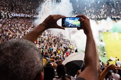 Torcedores nas arquibancadas filmam uma partida de futebol com um smartphone.